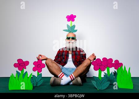 Bärtiger älterer Mann, der eine skurrile Yoga-Session genießt, umgeben von farbenfrohen Blumen in einer verspielten und kreativen Umgebung. Stockfoto