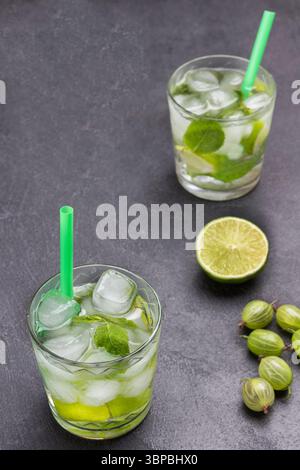 Zwei Gläser kaltes Getränk mit Limette, Minze und Eis. Strohhalm im Glas. Limettenscheiben und Stachelbeeren auf dem Tisch. Schwarzer Hintergrund. Draufsicht Stockfoto