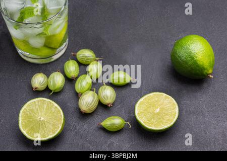 Limettenscheiben und Stachelbeeren auf dem Tisch. Zwei Gläser kaltes Getränk mit Limette, Minze und Eis. Schwarzer Hintergrund. Draufsicht Stockfoto