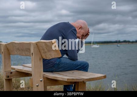 Mann, der auf einer Bank am Meer sitzt, mit dem Kopf in den Händen Stockfoto