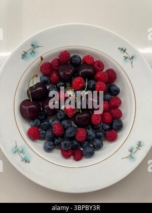 This image shows a top-down view of a white ceramic plate containing fresh cherries, blueberries, and raspberries. The fruits are arranged without spe Stockfoto