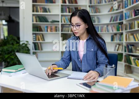 Eine junge asiatische Frau mit Brille studiert fleißig in einer Bibliothek, benutzt einen Laptop und macht Notizen. Stockfoto