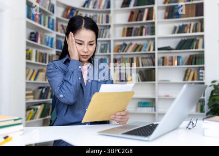 Eine asiatische Frau in einer Bibliothek ist sichtbar gestresst, wenn sie Dokumente hält und einen Laptop benutzt. Stockfoto