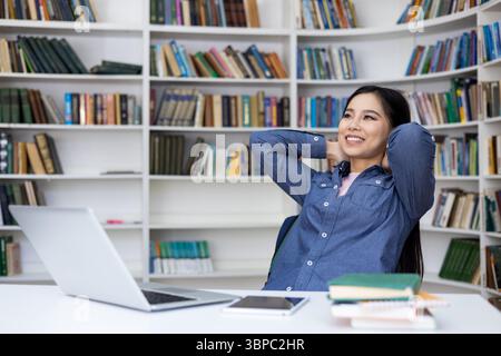 Eine junge asiatin entspannt sich in einer Bibliothek, lächelt mit den Händen hinter dem Kopf, umgeben von Büchern und einem Laptop. Stockfoto
