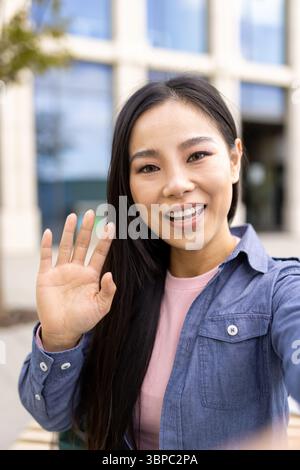 Eine asiatische Frau winkt hallo, während sie draußen ein Selfie macht, und lächelt warm in die Kamera. Stockfoto