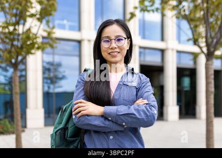 Eine lächelnde Asiatin steht selbstbewusst mit gekreuzten Armen, mit Brille und Rucksack vor einem modernen Gebäude. Stockfoto