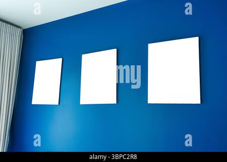 Drei leere, quadratische Leinwandrahmen, die auf einer tiefblauen Wand in einer Eigentumswohnung angeordnet sind. Moderne und minimalistische Ästhetik Stockfoto