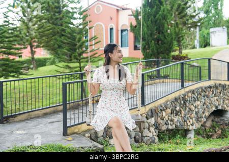 Glückliche asiatische Frau, die auf der Schaukel im tropischen Garten im Resort sitzt und lächelt Stockfoto