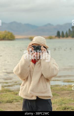 Junger asiatischer Tourist in gemütlicher Jacke, Hut genießt beim Fotografieren mit Retro-Kamera am Lake Tekapo im Herbst in Neuseeland Stockfoto
