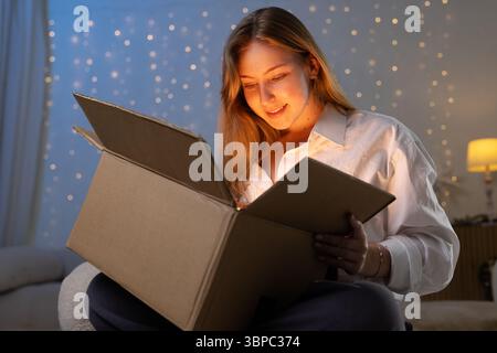 Die junge Frau öffnet zu Hause ein glühendes Paket mit fröhlichem Ausdruck, der magische Lieferung und festliche Überraschung symbolisiert Stockfoto