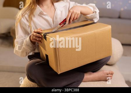 Nahaufnahme einer Frau, die zu Hause ein Paket mit einem Cutter öffnet, die E-Commerce-Zustellung und einen Lifestyle-Moment zeigt Stockfoto