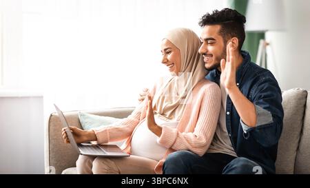 Fröhlich Schwanger Muslim Paar Mit Laptop Zu Hause Für Videoanruf Stockfoto
