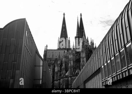Erleben Sie den Kölner Dom mit zwei gotischen Türmen, die sich über modernen Fassaden erheben, ein eindrucksvoller Dialog zwischen mittelalterlicher Architektur und zeitgenössischer Stadtform. Stockfoto