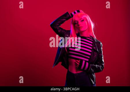 Stilvolle junge Frau mit langen Haaren, die unter dramatischer rosa und blauer Beleuchtung posiert. Das wunderschöne weibliche Modell mit Lederjacke und gestreiftem Crop-Top strahlt auffällige, trendige Mode und urbane Attitüde aus. Stockfoto