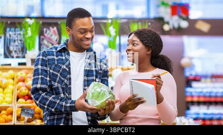 Einkaufen während des Coronavirus-Ausbruchs. Millennial schwarzes Paar Kauf frisches Gemüse, Überprüfung der Lebensmittelliste im Einkaufszentrum Stockfoto