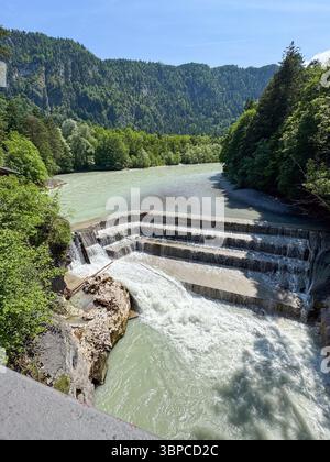 Der Fluss stürzt über einen gestuften Wasserfall, umgeben von üppig grünen Wäldern und felsiger Landschaft. Strahlend blauer Himmel über uns und sanfte Berge in der Ferne Stockfoto