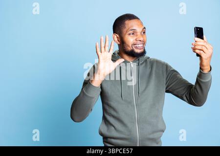 Ein afroamerikanischer Mann, der mit seiner Gruppe von Freunden online telefoniert und über eine Internet-Videokonferenz im Studio plaudert und tratscht. Fröhlicher junger Kerl steht vor blauem Hintergrund. Stockfoto
