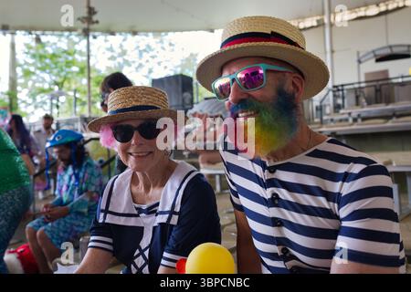 Zwei ältere Erwachsene, ein Mann mit Regenbogenbart und eine Frau mit rosa Haaren, sitzen draußen und tragen Strohhüte und Sonnenbrille. Sie sind in nautischen... Stockfoto