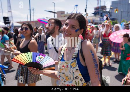 Eine junge Frau mit blauem Glitzer an Arm und Gesicht hält einen Regenbogen-Handfan bei einem lebhaften Outdoor-Event. Sie trägt einen gemusterten Jumpsuit. Andere in Stockfoto