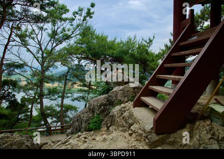 Buyeo County, Südkorea - 13. Juni 2025: Eine Reihe steiler Holztreppen führt hinauf zum Pavillon auf dem Nakwaam Rock, umgeben von Kiefern und darüber Stockfoto