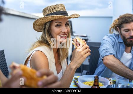 Glückliche Frau, die Hot Dog auf einer Grillparty auf dem Dach mit Freunden isst und gutes Essen und Gesellschaft genießt Stockfoto