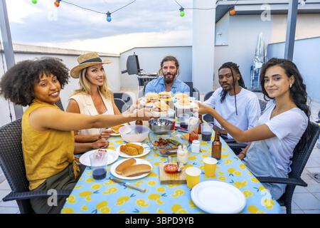 Verschiedene Freunde lachen und genießen köstliche Burger, während sie den Sommer auf einer lebhaften Grillparty auf dem Dach feiern Stockfoto