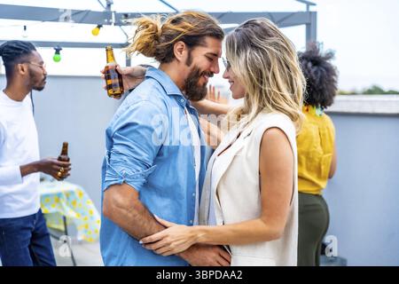 Glückliches Paar, das sich zärtlich auf einer Dachparty umgibt, umgeben von Freunden, die Drinks und gute Gesellschaft genießen Stockfoto