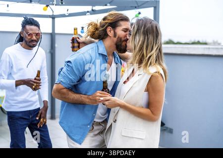 Junge Paare küssen und Hände halten auf der Dachparty, während Freunde Bier trinken und Spaß haben Stockfoto