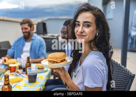 Junge Frau, die einen köstlichen Burger genießt, während sie mit Freunden auf einer lebhaften Grillparty auf dem Dach feiert und die Sommerstimmung genießt Stockfoto
