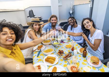 Fröhliche Multi-ethnische Freunde genießen eine Grillparty auf dem Dach, machen Selfie und toasten mit Getränken Stockfoto