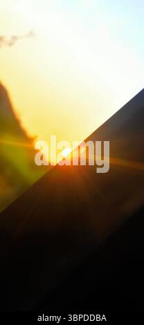 Kreativer Blick auf die untergehende Sonne, die teilweise durch ein diagonales Glas blockiert wird und bei Sonnenuntergang warme Sonnenstrahlen durch den goldenen Himmel wirft. Stockfoto