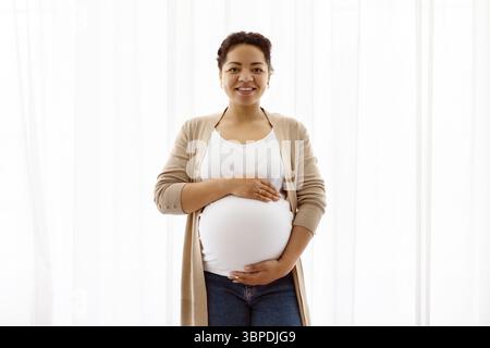 Lächelnde schwangere Frau, die ihren Bauch umgibt und selbstbewusst in die Kamera schaut Stockfoto