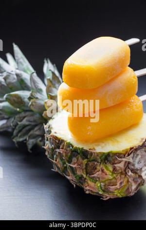 Drei lebendige Mango-Ananas-Eis-Pops auf einer frischen Ananashälfte präsentieren ein farbenfrohes und erfrischendes Dessert, ideal für heiße, sonnige Tage, perfekt für ein Sommertreffen. Stockfoto