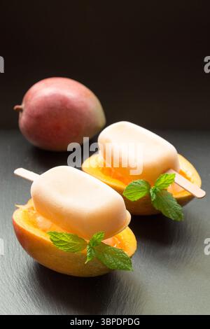 Mangosorbet-Eis auf halb geschnittenen Mangobrüchten hinter Mango auf schwarzem Hintergrund. Mangobruchtsorbet-Eis-Eis. Vertikal. Stockfoto
