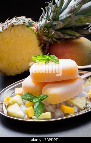 Exotisches Sorbeteis auf Silberplatte mit Eiswürfeln gehackte Ananasmangobiteile, Minze nahe der Hälfte der Ananas, Mango auf schwarzem Hintergrund. Ananas-Mangosorbet-Eis-Eis. Stockfoto