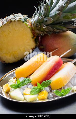 Ananas-Mango-Erdbeersorbet-Eis auf Silberplatte mit Eiswürfeln, Fruchtstücke nahe der Hälfte der Ananas, Mango auf schwarzem Hintergrund. Ananasmangoherdbeersorbet-Eis-Eis-Eis. Stockfoto