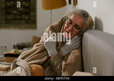 Ältere kaukasische Frau sitzt drinnen, in Decke gehüllt und lächelt friedlich Stockfoto