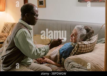 Senior Black man füttert ältere kaukasische Frau, die zu Hause auf dem Sofa liegt Stockfoto