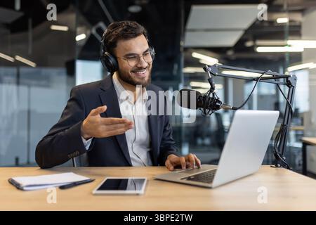 Ein Geschäftsmann im Anzug lächelt beim Podcast, trägt Kopfhörer, benutzt einen Laptop und spricht in einem modernen Büroumfeld in ein Mikrofon. Stockfoto