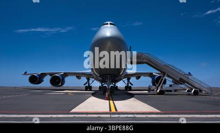Die Queen, ein B747 großes Verkehrsflugzeug, von vorne gesehen, mit angebrachter Einstiegtreppe, parkt auf dem Flughafengelände unter klarem blauem Himmel. Stockfoto