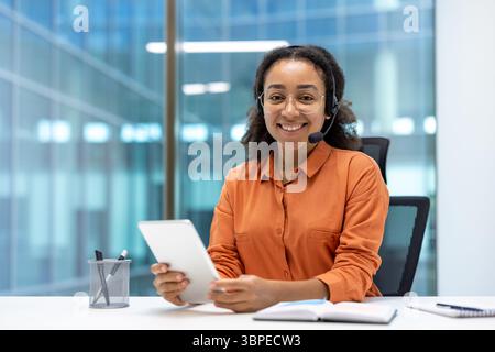 Eine lächelnde Frau, die ein Headset und eine Brille trägt und ein Tablet in einer modernen Büroumgebung hält. Sie blickt direkt in die Kamera. Stockfoto