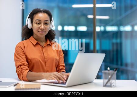 Eine lächelnde Frau mit Brille und Kopfhörern sitzt an einem Schreibtisch und arbeitet an einem Laptop in einer modernen Büroumgebung. Stockfoto