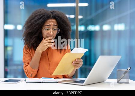 Eine besorgte junge Frau mit Brille untersucht Dokumente, während sie an ihrem Schreibtisch arbeitet, und sieht gestresst aus. Stockfoto