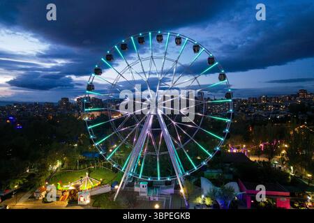 Ein Riesenrad, das in der Abenddämmerung im Victory Park, Jerewan, Armenien, mit Blick auf die Skyline der Stadt und die Attraktionen des Vergnügungsparks leuchtet. Stockfoto