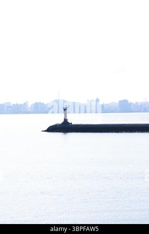 Ruhige Küstenszene mit einem Leuchtturm an einem ruhigen Tag inmitten eines sanften Hintergrunds der Skyline der Stadt und sanftem Wasser, das das Licht reflektiert Stockfoto