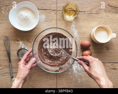 Weibliche Hände bereiten Schokoladenschwammkuchen vor. Zutaten auf einem Holztisch Stockfoto