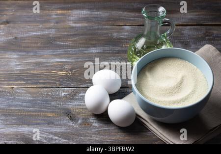 Grieß in der Schüssel, Eier und Pflanzenöl auf rustikalem Holztisch, Zutaten zum hausgemachten Kochen und Backen Stockfoto