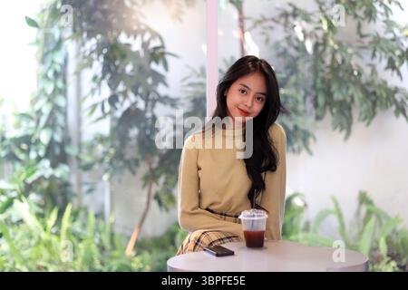 Attraktive junge asiatische Frau lächelt hinter einer Tasse Eiskaffee Americano auf einem weißen Tisch. Ein Porträt einer wunderschönen indonesischen Teenagerin Ssmil Stockfoto