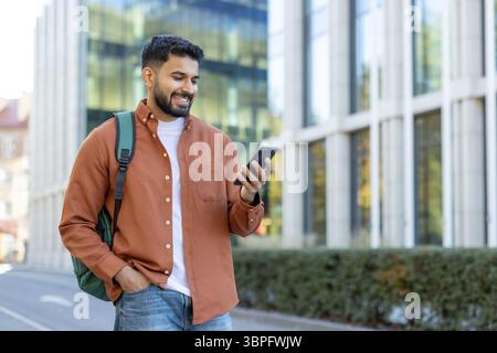 Ein lächelnder Student, der draußen sein Handy benutzt, während er vor einem modernen Gebäude läuft Stockfoto