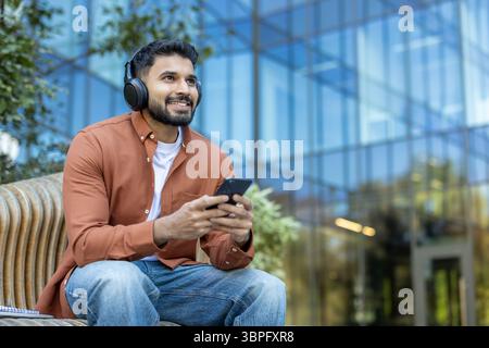 Ein lächelnder Mann mit Kopfhörern benutzt sein Telefon, während er draußen sitzt, in der Nähe eines modernen Gebäudes. Stockfoto